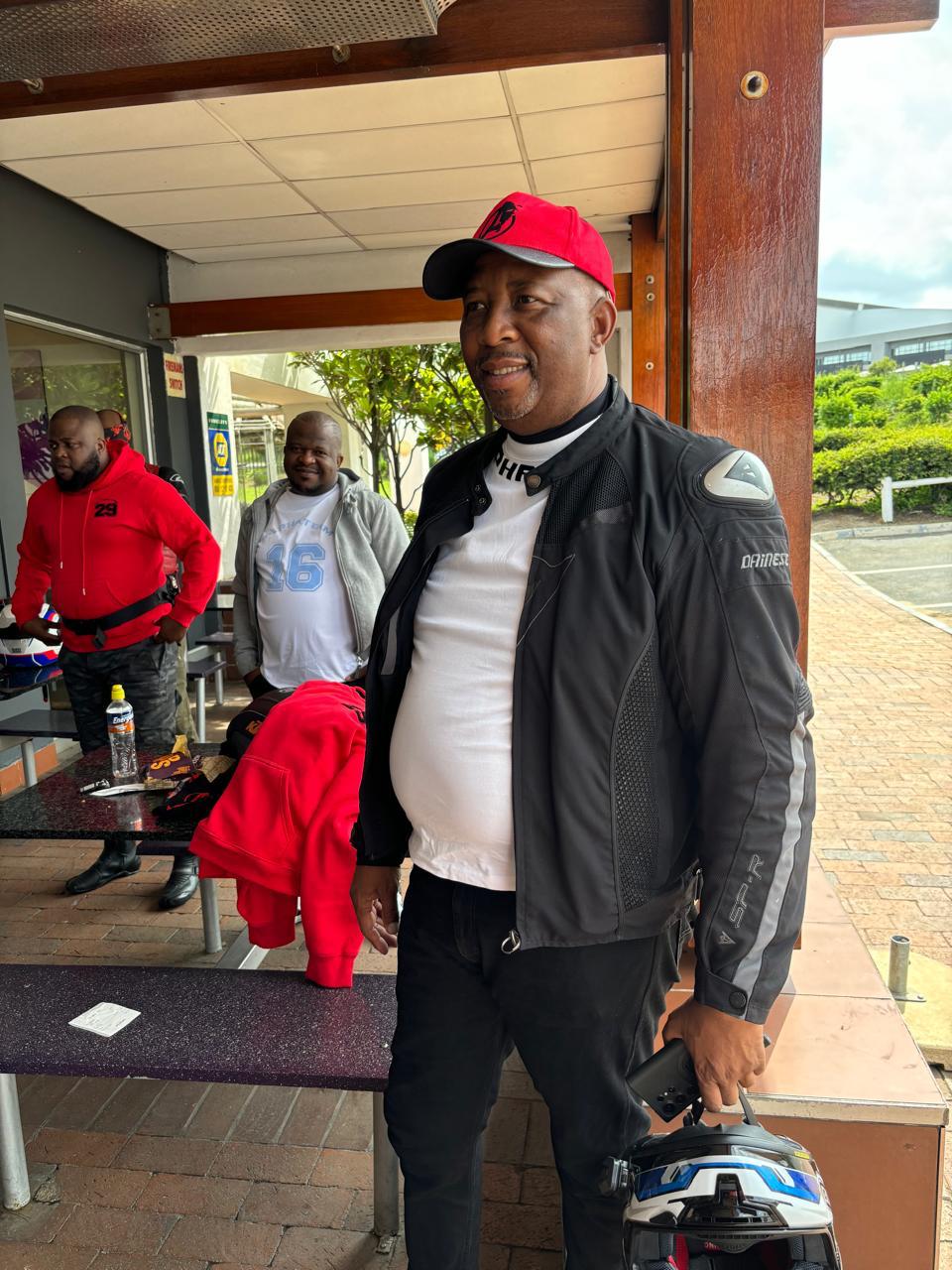 Alpha Team rider in a red club cap and Dainese jacket holding his helmet